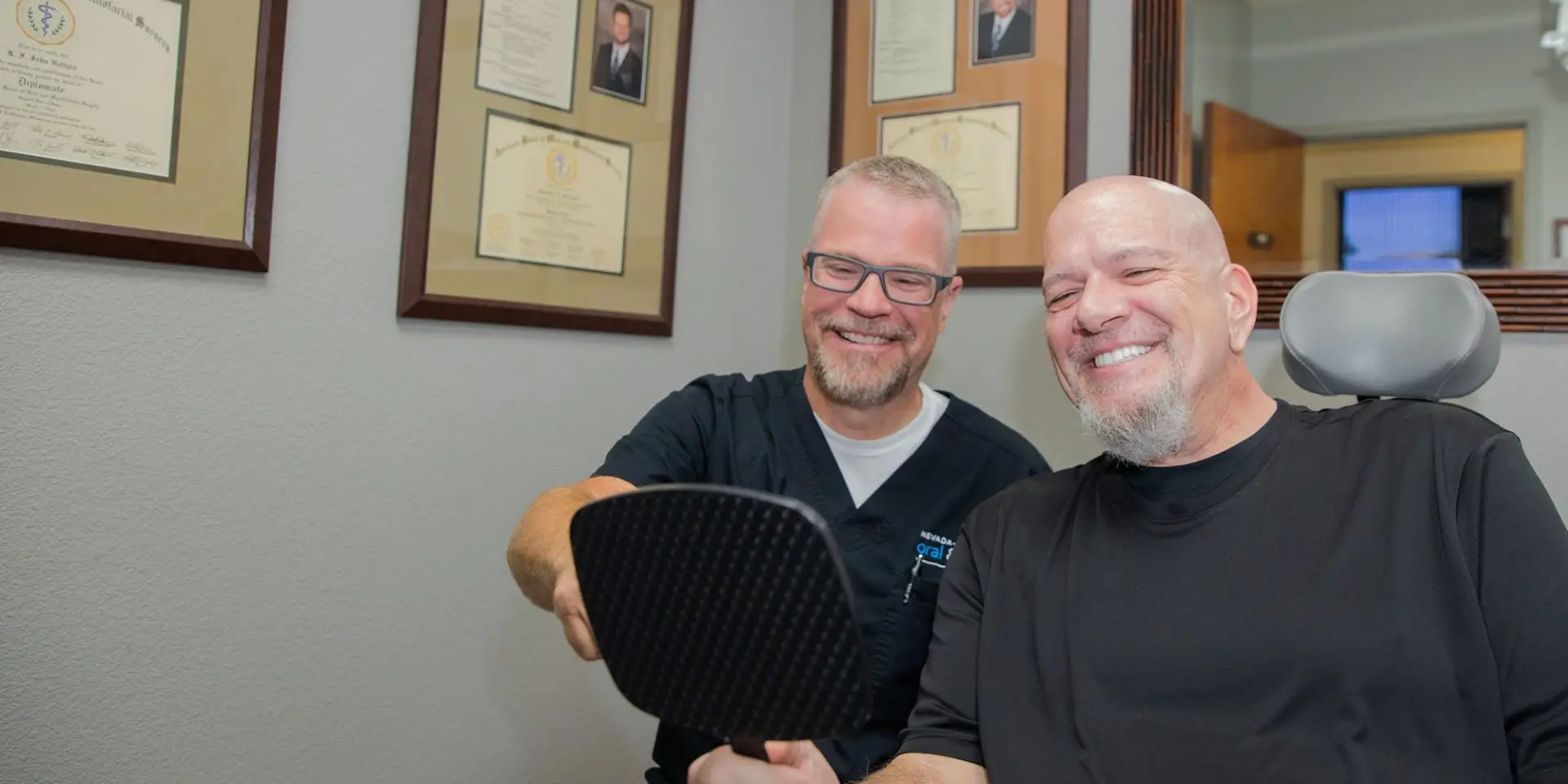 Two men sit smiling, one holds a mirror. Certificates and framed photos hang on the wall in a professional office setting.