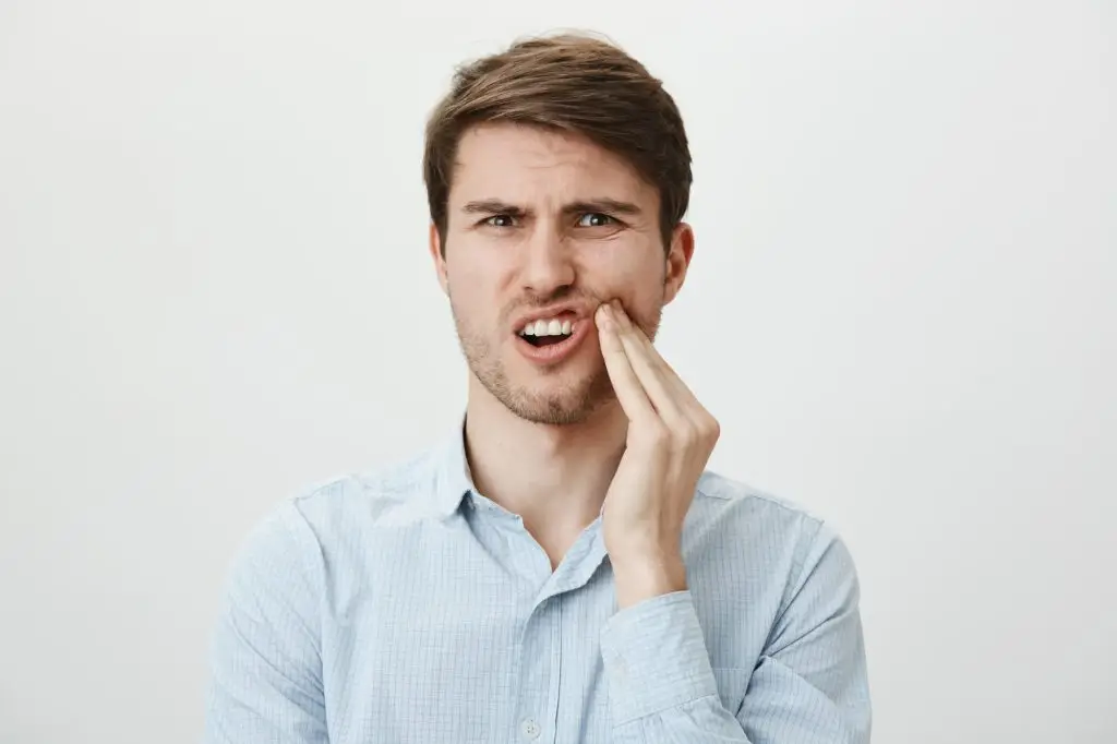 A man in a light blue shirt holds his cheek with a pained expression, suggesting toothache, against a plain white background.