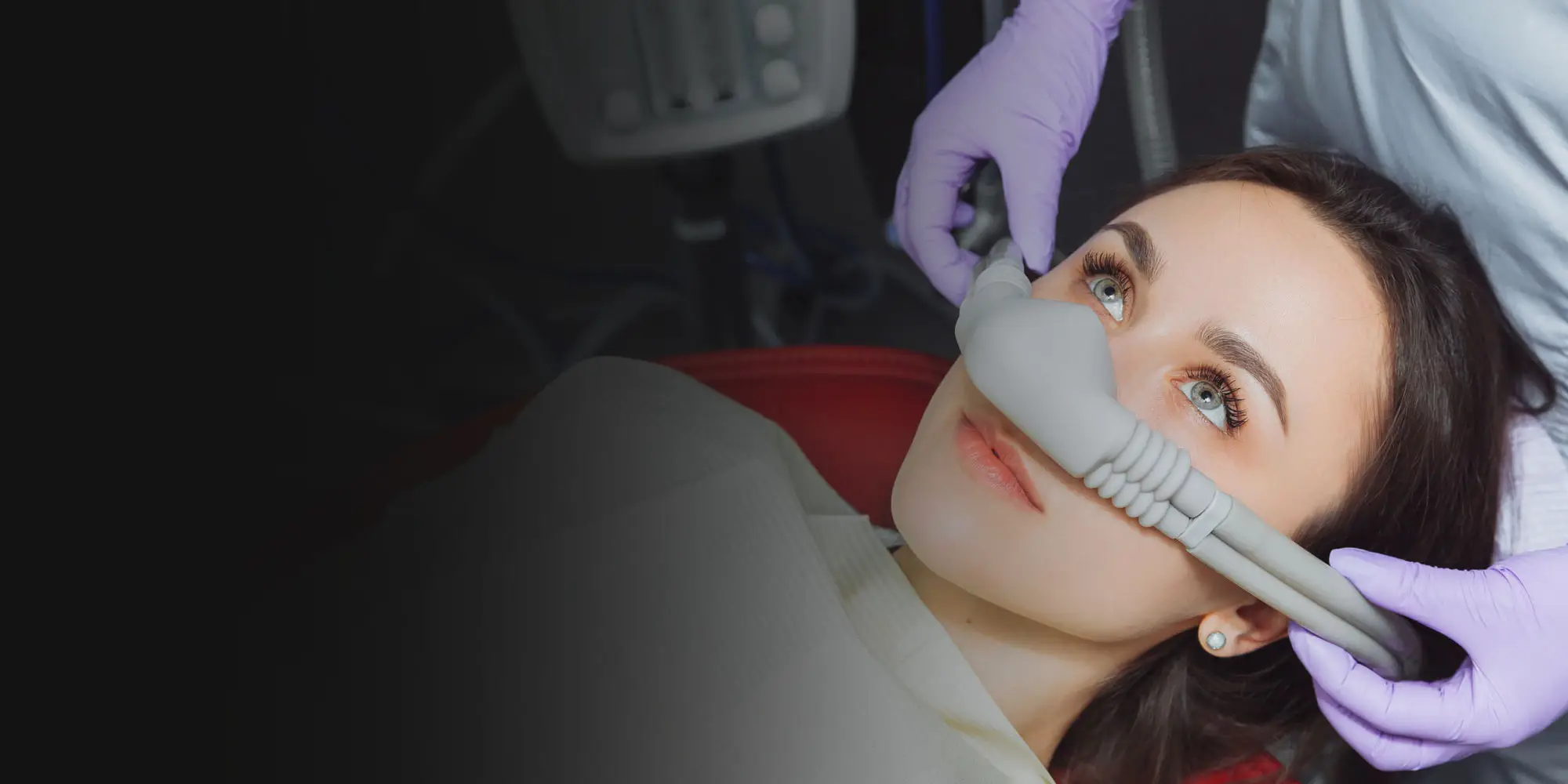 A person lies on a dental chair, wearing a nitrous oxide mask administered by someone with purple gloves. The background shows dental equipment in a clinical setting.