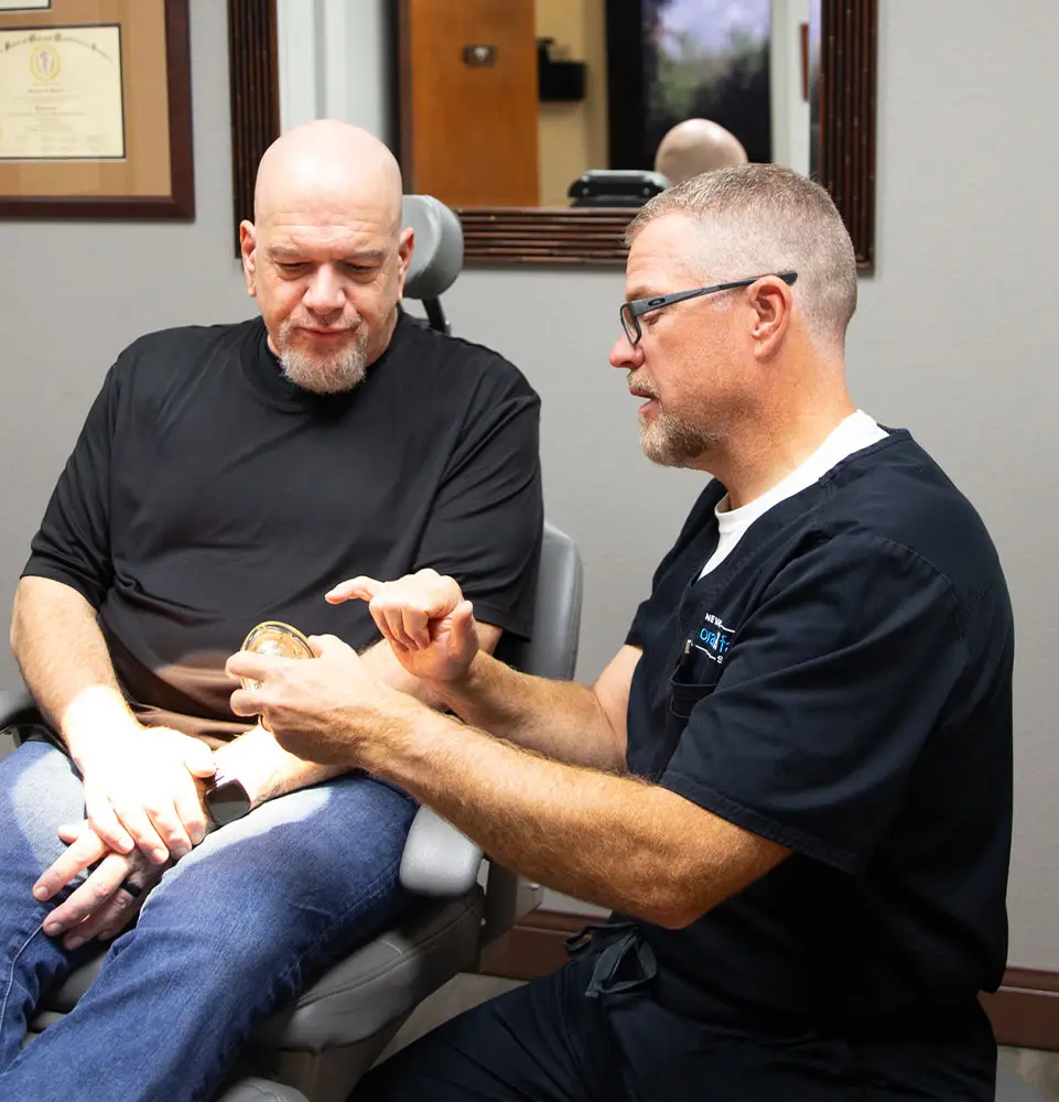 A doctor explains a medical device to a seated patient, in a clinic setting with framed certificates on the wall.