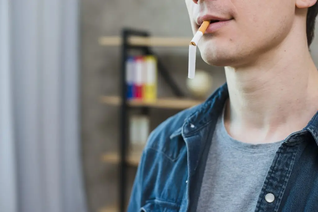 Person holds an unlit cigarette in their mouth, wearing a denim shirt over a gray T-shirt. A blurred background features a bookshelf with books and a globe.