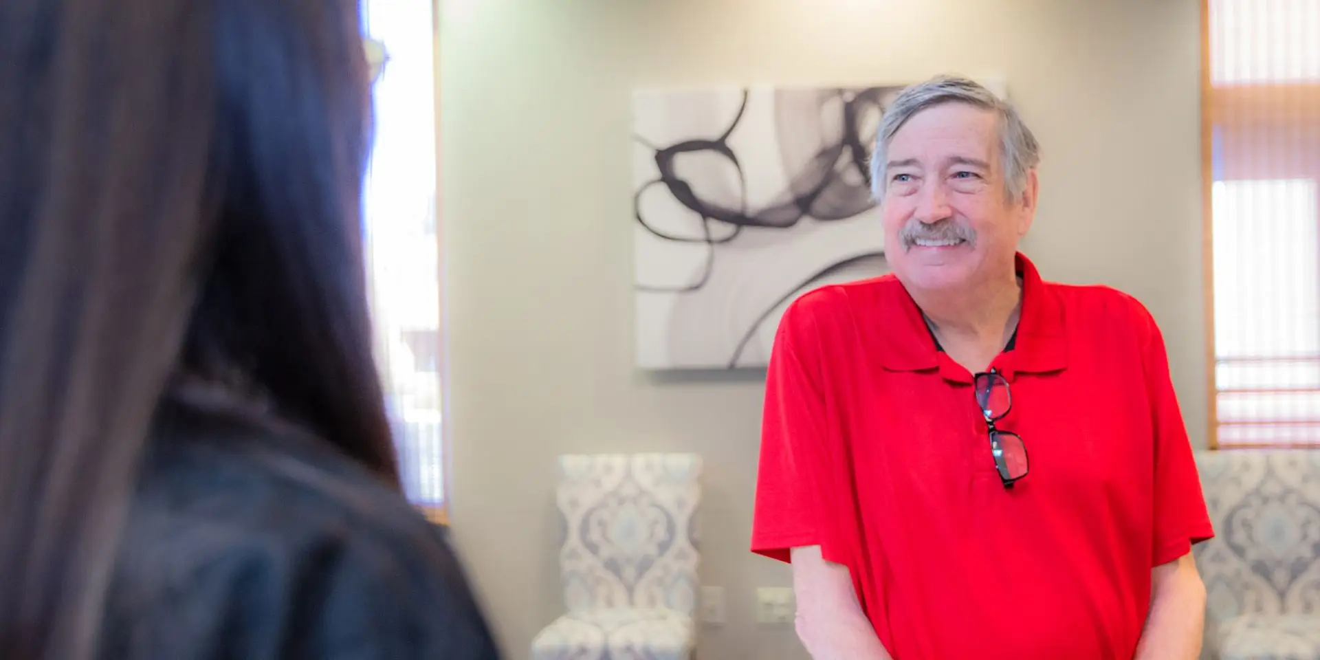 A man in a red shirt smiles while standing at a reception desk, interacting with another person. The setting is an office with abstract wall art and patterned chairs.