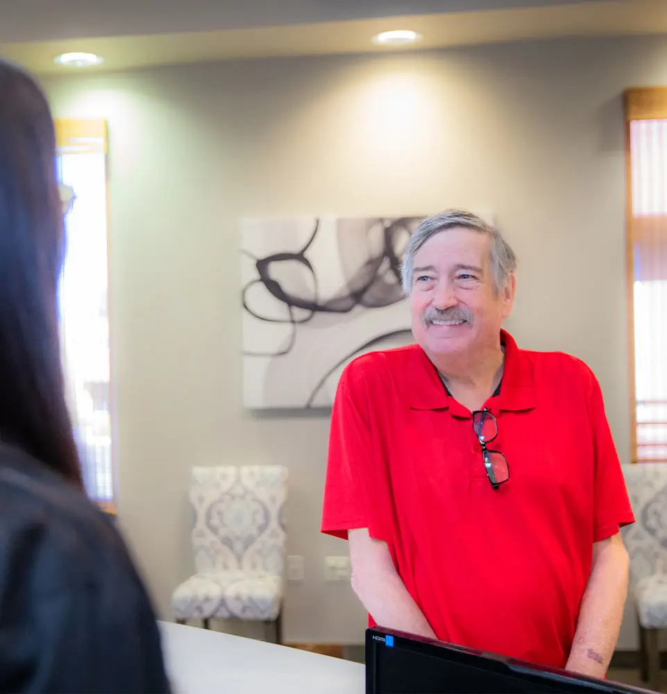 A man in a red shirt smiles while standing at a reception desk, interacting with another person. The setting is an office with abstract wall art and patterned chairs.