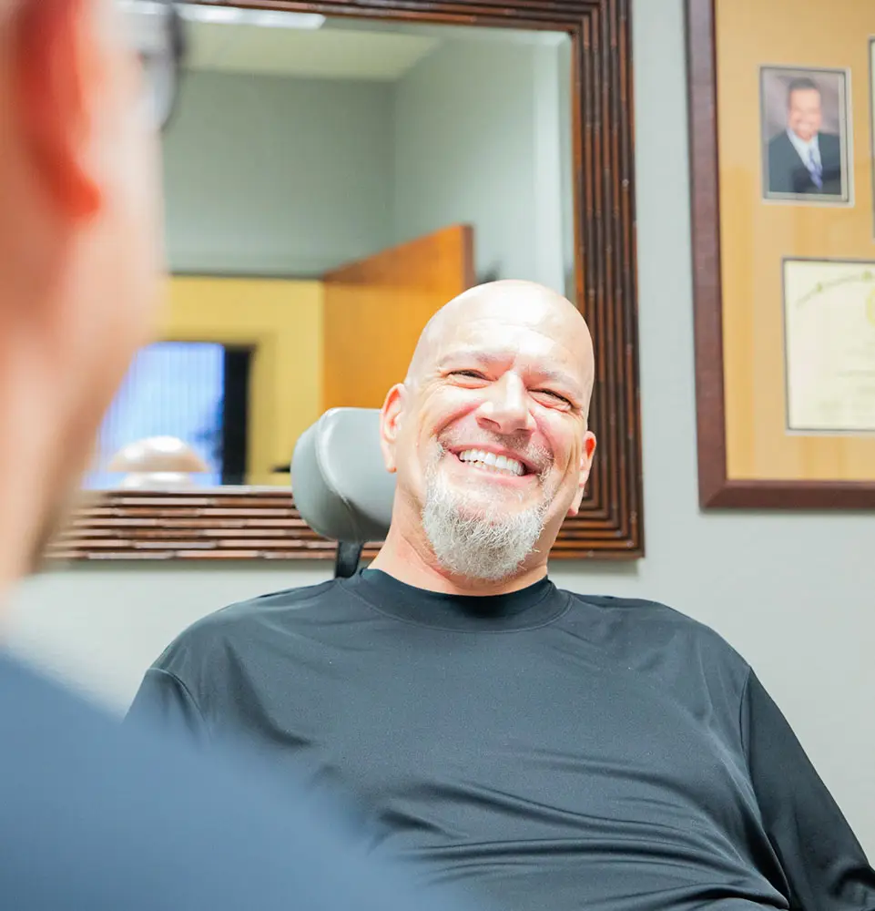 A man with a goatee smiles while sitting in a chair, facing another person. Framed certificates and a photo decorate the office wall.