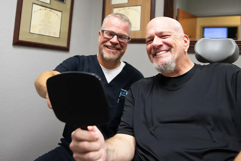 Two men smile while sitting, one holding a mirror. They appear in a professional setting, with framed certificates on the wall in the background.