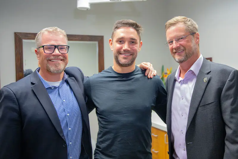 Three men stand closely with arms around each other, smiling warmly. They are in a room with light-colored walls, a mirror, and a wooden cabinet visible in the background.