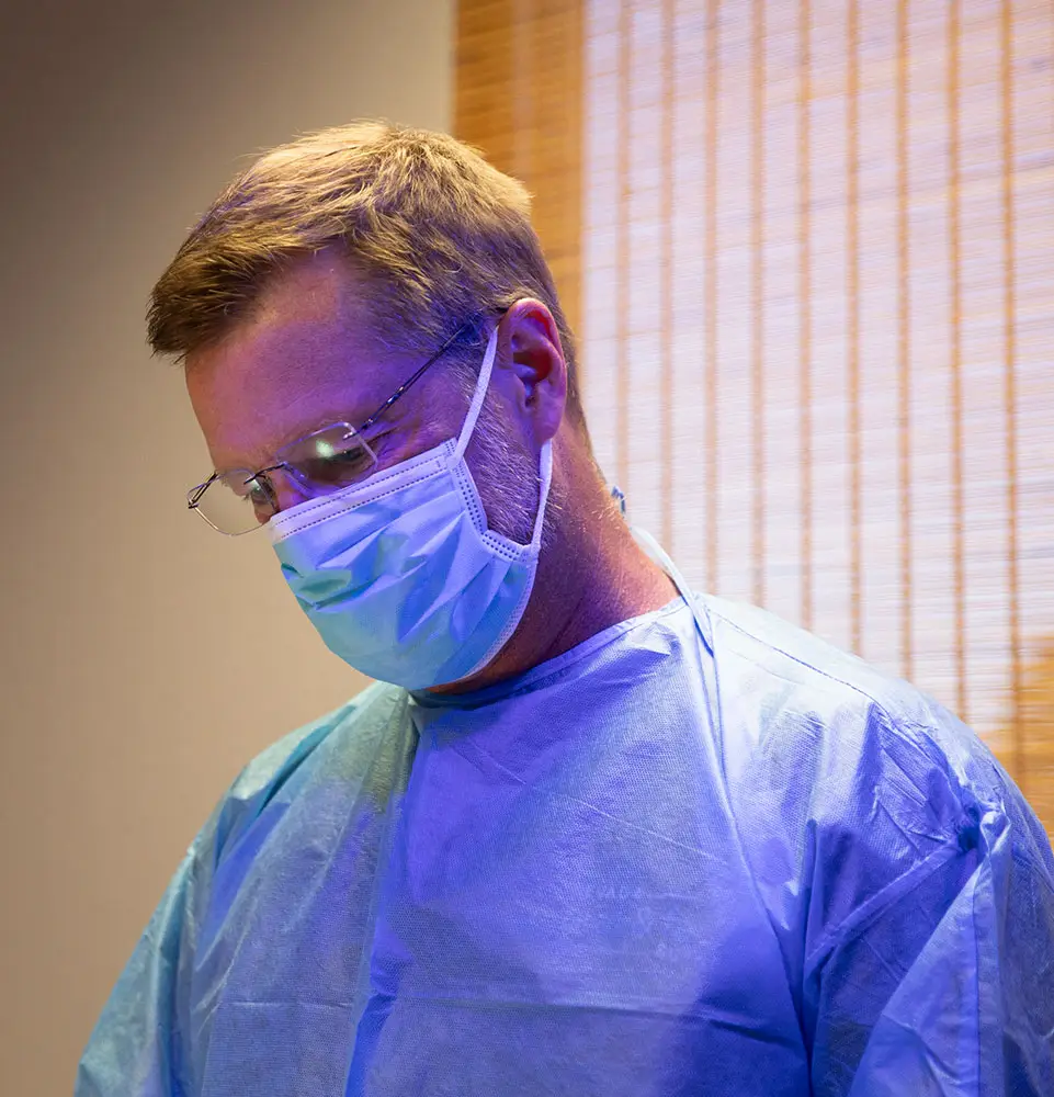 A person wearing a surgical mask and gown looks downward, standing in a softly illuminated room with subtle, warm lighting and vertical blinds in the background.