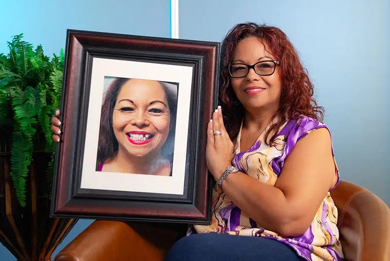 A woman, seated in a chair, holds a framed photo of herself smiling. She's dressed in a colorful top, against a light blue background with potted ferns nearby.