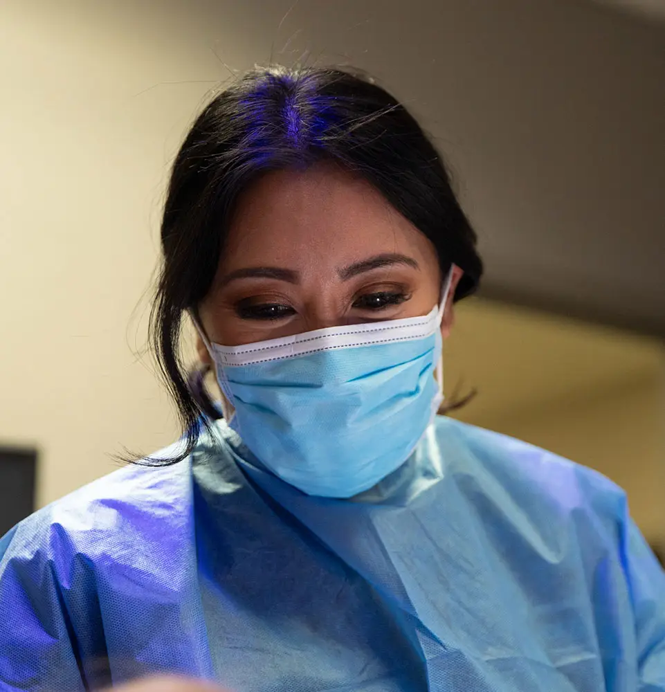 A person wearing a blue surgical mask and gown leans forward attentively in a clinical setting, under a soft light.