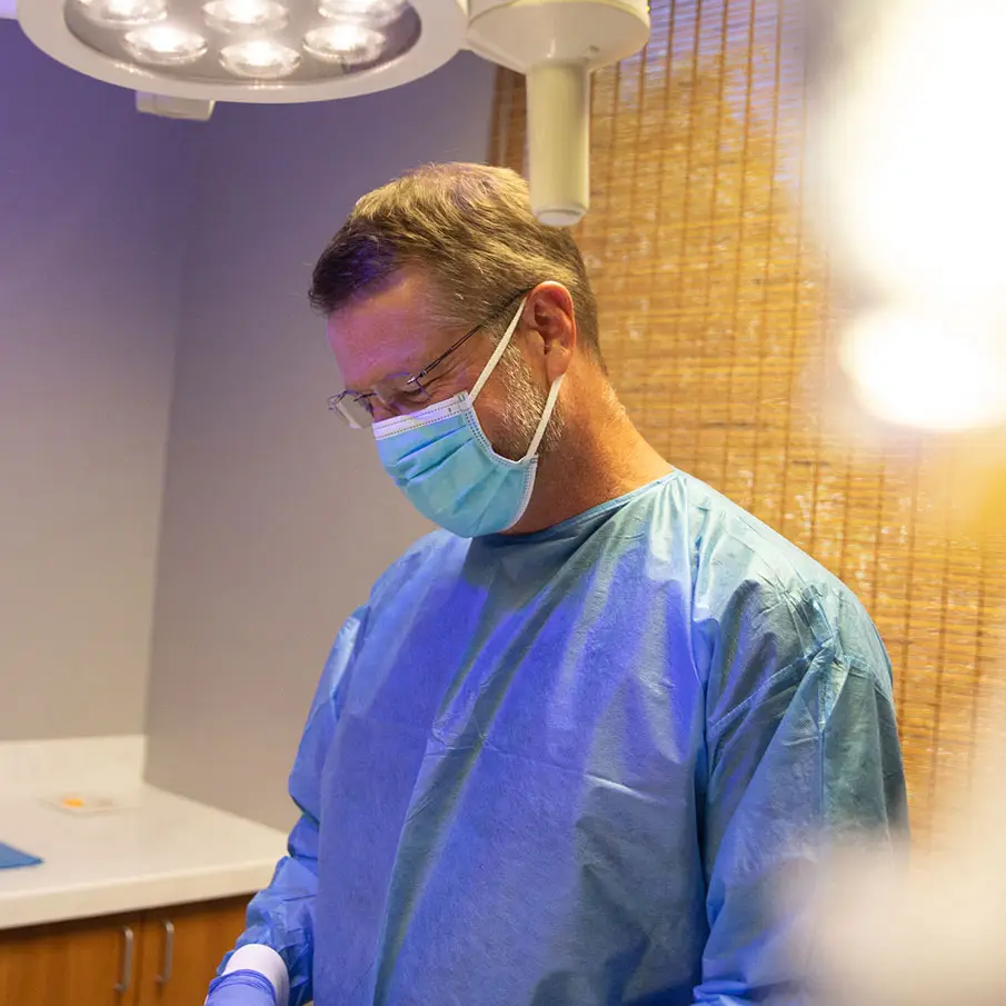 A medical professional wearing a mask and surgical scrubs focuses intently, illuminated by overhead lights, in a clinical room with wooden privacy screens and white cabinets.