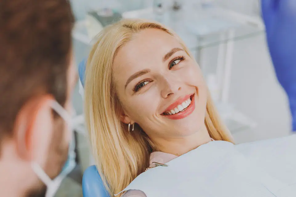 A woman smiles while reclining in a dental chair, conversing with a dentist in a medical setting.