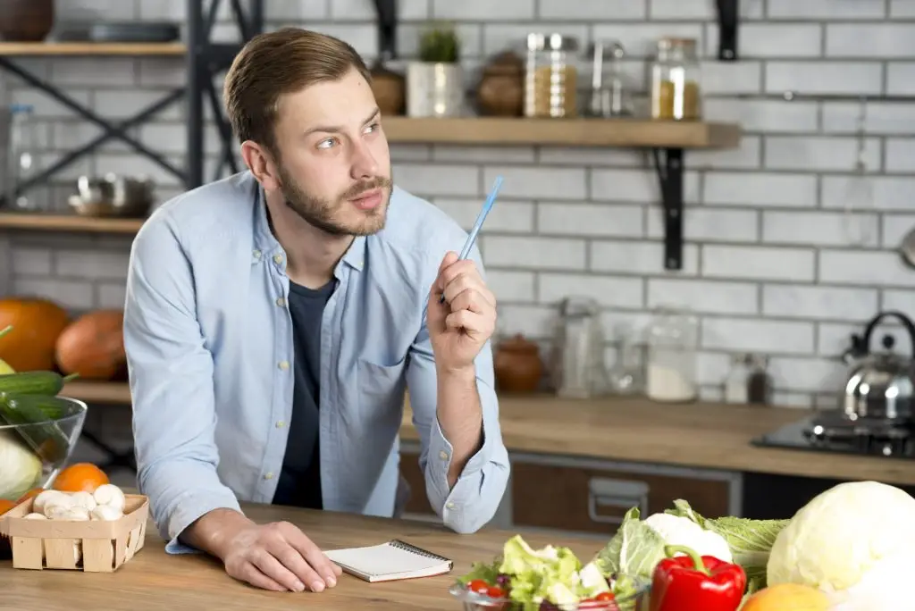 A man leans on a kitchen counter, thoughtful, holding a blue pen over a notepad thinking about a recipe to write down. Vegetables like bell peppers and cabbages are spread around. Shelves and kitchenware fill the background.