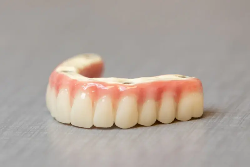 Dentures are positioned on a flat surface, displaying a row of artificial teeth embedded in a pink gum-like base, with a neutral-colored background.